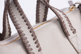 Dark gray whipstitching accents on satchel straps. Gold zipper on a cream colored bag. 